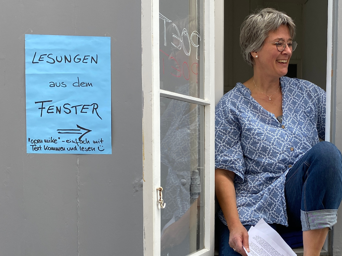 HannaH Rau sitzt lachend im Fenster der WortWerft, daneben ein handgeschriebenes Schild: Lesungen aus dem Fenster – open mike, einfach mit Text kommen und lesen